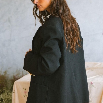 Mulher usando casaco preto com bolsos grandes em fundo neutro e tecido com flores.