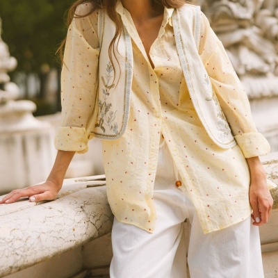 Camisa amarela com pequenas estampas, colete branco bordado e calças brancas num cenário exterior.