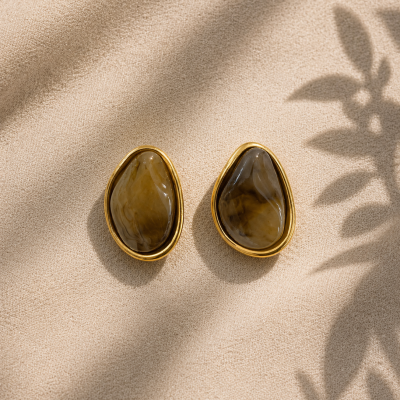 Brincos com pedra castanha e moldura dourada sobre fundo bege com sombras de folhas
