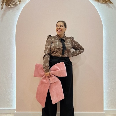 Mulher com camisa castanha estampada, calças pretas largas, segurando grande laço rosa, em fundo claro com arco e folhas secas.