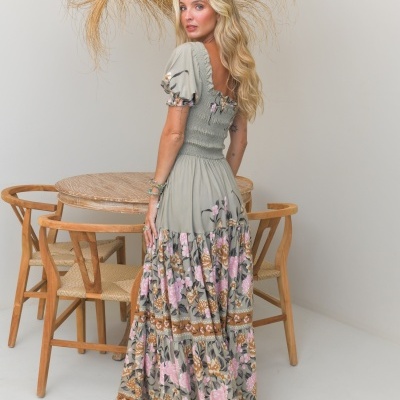 Vestido longo verde com flores rosas, modelo em ambiente minimalista com mesa e cadeiras de madeira