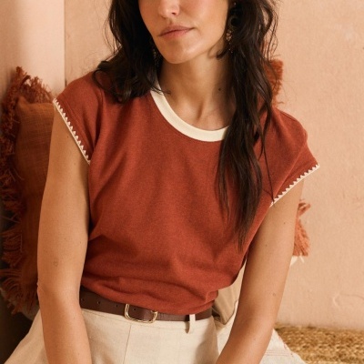 Mulher com t-shirt ferrugem e calças bege sentada num interior com almofada e parede cor de pêssego.