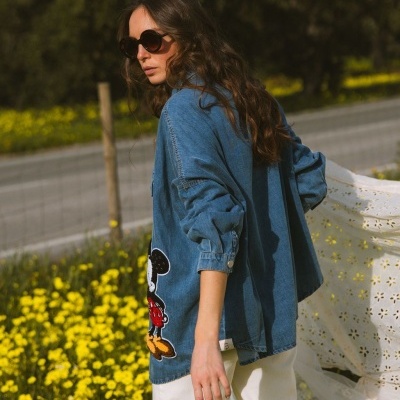 Mulher a usar camisola de ganga azul com padrão do Mickey Mouse e óculos de sol num campo de flores amarelas.