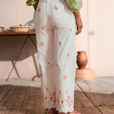 Calças brancas bordadas com flores cor-de-rosa e sandálias rosa num ambiente rústico.