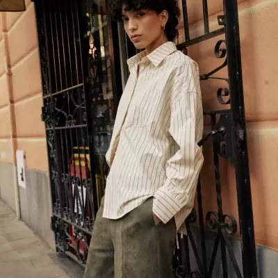 Pessoa com camisa riscada branca e calções verdes encostada a portão metálico preto