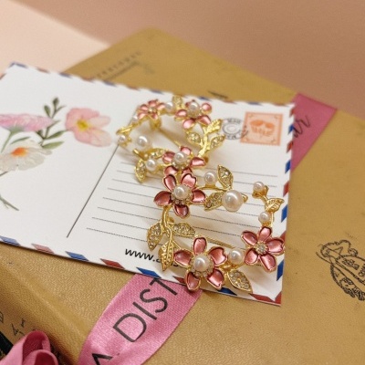 Broche floral dourado com pérolas numa embalagem com cartão postal e fita rosa.