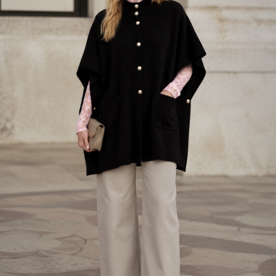 Mulher com poncho preto, camisa rosa de renda, calças bege largas e carteira bege