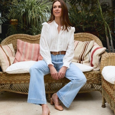 Mulher sentada num sofá de verga com almofadas bege e rosas, veste camisa branca, jeans azuis e sapatilhas castanhas.