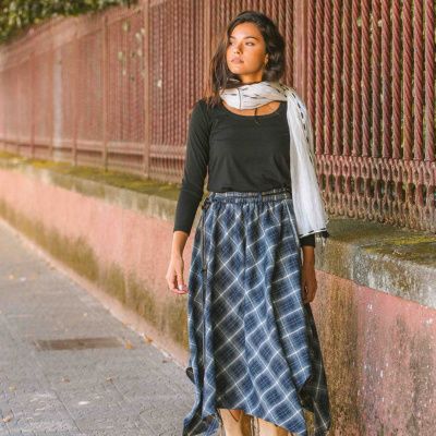 Mulher com blusa preta, saia xadrez azul, botas bege e lenço branco a passear junto a vedação vermelha