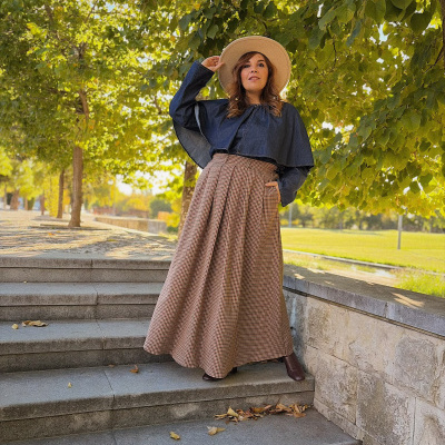 Mulher com blusa denim azul-escura, saia xadrez castanha e bege, chapéu bege e botas castanhas em escadaria ao ar livre com árvores verdes.