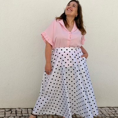 Mulher de blusa rosa e saia branca com bolinhas pretas junto a parede branca.