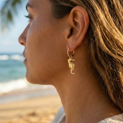 Brinco de argola dourada com pendente em forma de cavalo-marinho numa orelha feminina na praia
