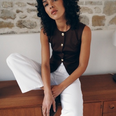 Mulher com top castanho e calças brancas sentada numa cómoda de madeira com parede de pedra ao fundo