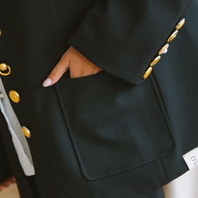 Blazer preto com botões dourados e bolso frontal, mão no bolso, etiqueta maïtté