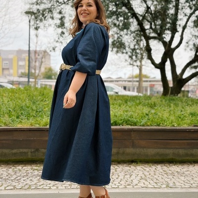 Mulher vestindo vestido azul escuro com cinto bege e sandálias castanhas, em ambiente exterior com árvores e pavimento de pedra.
