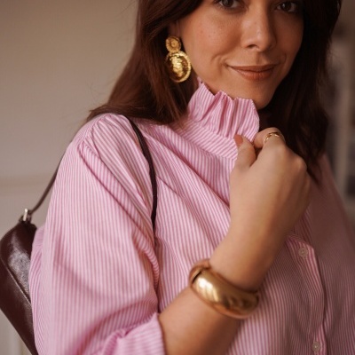 Mulher com blusa rosa riscada branca, acessórios dourados e mala castanha ao ombro