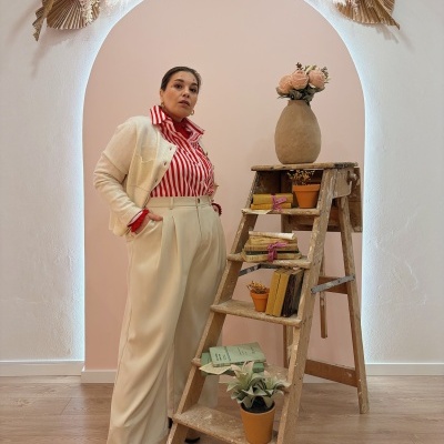 Mulher vestida com camisa vermelha de riscas, casaco e calças beges, sapatos castanhos junto a escada de madeira com plantas e livros, parede rosa claro com arco e decoração seca.