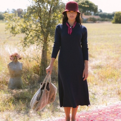 Mulher com vestido azul escuro, cachecol vermelho, chapéu vinho e bolsa xadrez em campo com tapete vermelho.