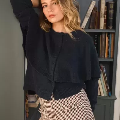 Mulher com casaco azul escuro e calças xadrez junto a estante de livros