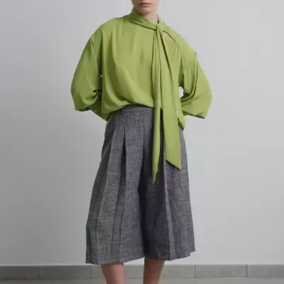 Mulher com blusa verde e calças culotte cinzentas num fundo interior cinzento