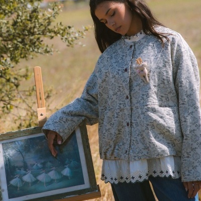 Mulher vestindo casaco floral cinza e branco com broche de rena, blusa branca rendada e calças jeans azuis, ao ar livre junto a uma moldura com pintura de bailarinas.