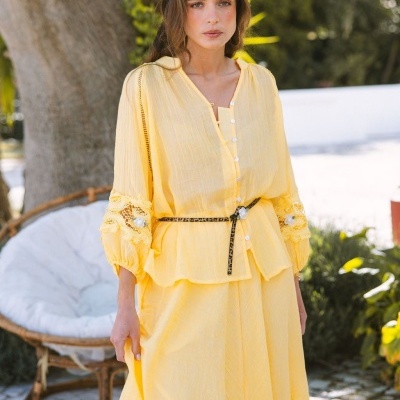 Mulher com conjunto amarelo, blusa com botões e renda nas mangas, cinto animal print, ao ar livre