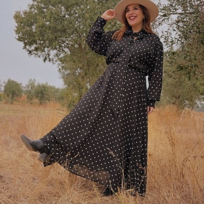 Mulher a usar vestido comprido preto com bolinhas brancas e chapéu bege num campo seco