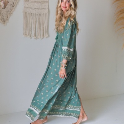 Mulher com vestido comprido verde estampado, descalça, em ambiente interior com decoração de macramé