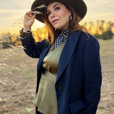Mulher com casaco azul escuro, camisa verde, lenço preto e branco e chapéu castanho ao ar livre ao pôr do sol