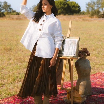 Mulher com camisa branca, saia plissada castanha e botas bege ao ar livre sobre tapete vermelho com cavalete de pintura e busto