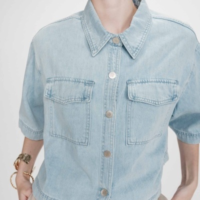 Camisa de ganga azul clara com botões e bolsos frontais em fundo branco.