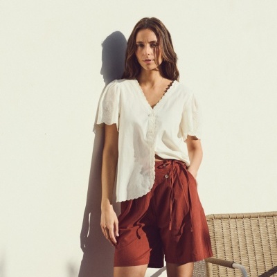 Mulher de pé com blusa branca e calções cor ferrugem junto a cadeira de vime
