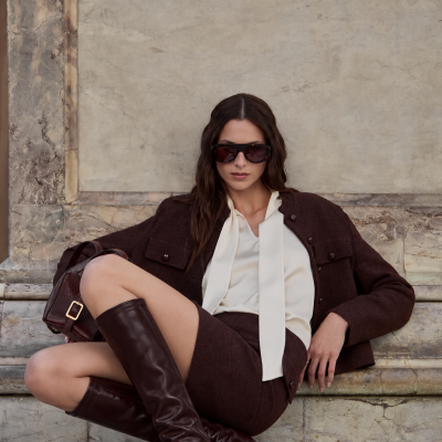 Mulher com roupa castanha e bege sentada contra parede de pedra.