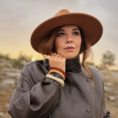 Mulher com casaco castanho escuro, chapéu castanho claro e bijoux em ambiente exterior