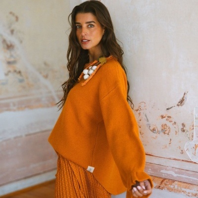 Mulher com camisola de malha laranja e saia plissada com padrão de pontos brancos, contra parede texturada