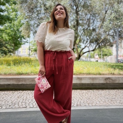 Mulher com blusa branca bordada, calças vermelhas largas, sandálias castanhas e clutch rosa floral ao ar livre