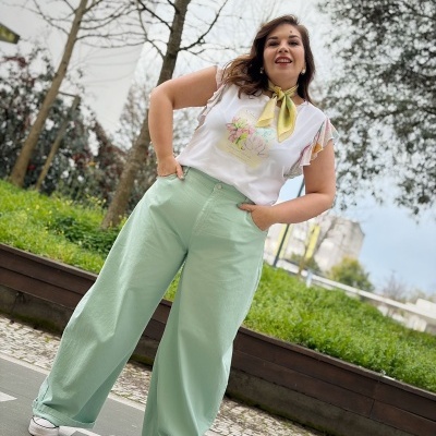 Pessoa com calças largas verdes claras, camisola branca com estampado floral, lenço amarelo e sapatos brancos ao ar livre