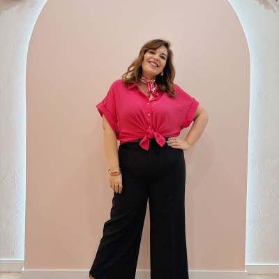 Mulher sorridente vestindo camisa rosa, calças pretas largas e botas camel em ambiente decorado com arco