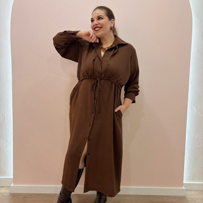 Vestido comprido castanho com botões e cinto, botas escuras, parede rosada com arco branco ao fundo