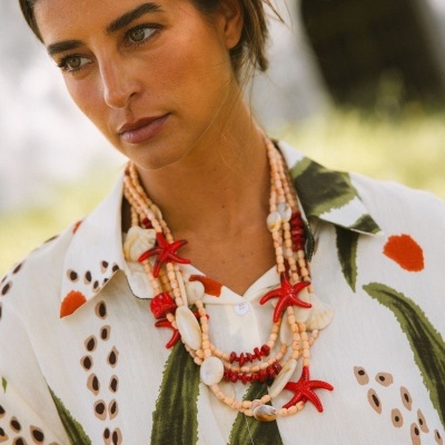 Colar coral e conchas em camisa estampada creme com folhas verdes e manchas laranja e pretas.