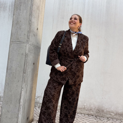 Mulher com fato castanho floral, camisola branca, botins castanhos, cachecol colorido e mochila preta numa rua com parede branca e coluna de betão.