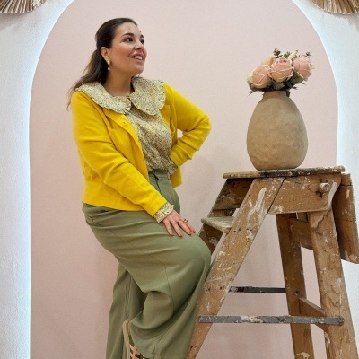 Mulher vestindo blusa amarela e saia verde junto a escada de madeira com vaso e flores