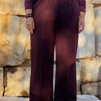 Calças largas vinho com cós elástico e blusa rendada roxa, usadas com acessórios dourados e pulseira roxa, fundo de pedra.