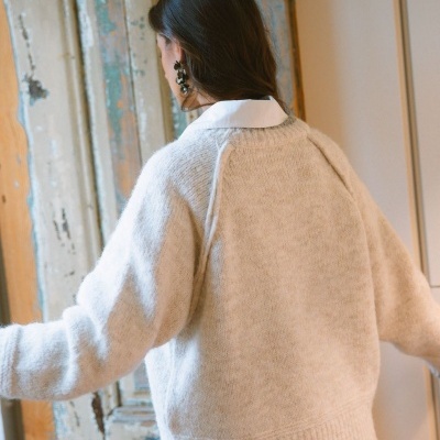 Mulher de costas com camisola de malha bege, camisa branca e brincos pretos, parede e porta antigas no fundo.