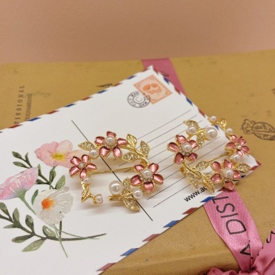 Broches dourados com flores cor-de-rosa e pérolas sobre postal floral e livro com laço rosa.