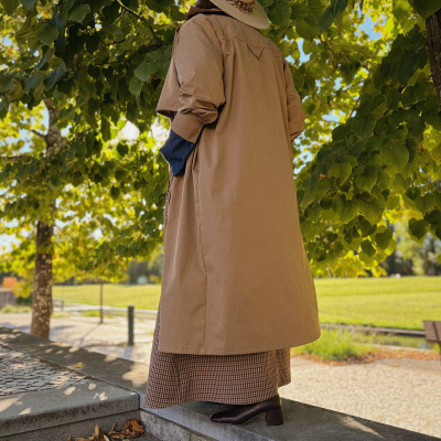 Pessoa vista de costas com casaco bege, vestido xadrez castanho, botas castanhas e chapéu bege num parque com árvores.
