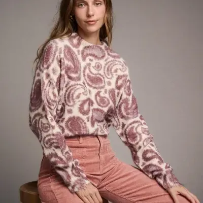 Mulher com camisola de padrão paisley creme e rosa e calças cor de rosa sentada num banco de madeira.