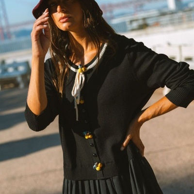 Mulher com cardigan preto com botões decorativos, vestido preto plissado e chapéu vinho ao ar livre