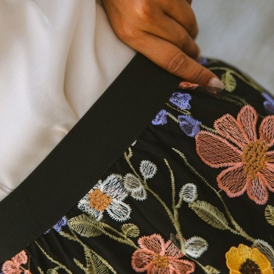 Saia preta com bordados florais coloridos e cinta elástica, com mão pousada ao lado sobre tecido branco.