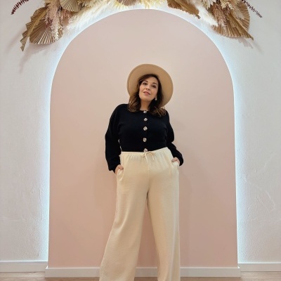 Mulher com cardigan preto e calças bege em parede rosa com arco branco e plantas secas